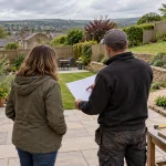 A gardener and homeowner compare a blank project sheet and paving samples beside a sloped garden.