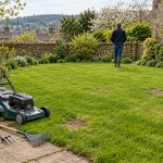 A simple mower and hand tools beside a lawn with a few bare patches in a modest garden.