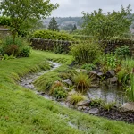 A planted rain garden and shallow grass-lined channel managing runoff beside a West Yorkshire stone home.